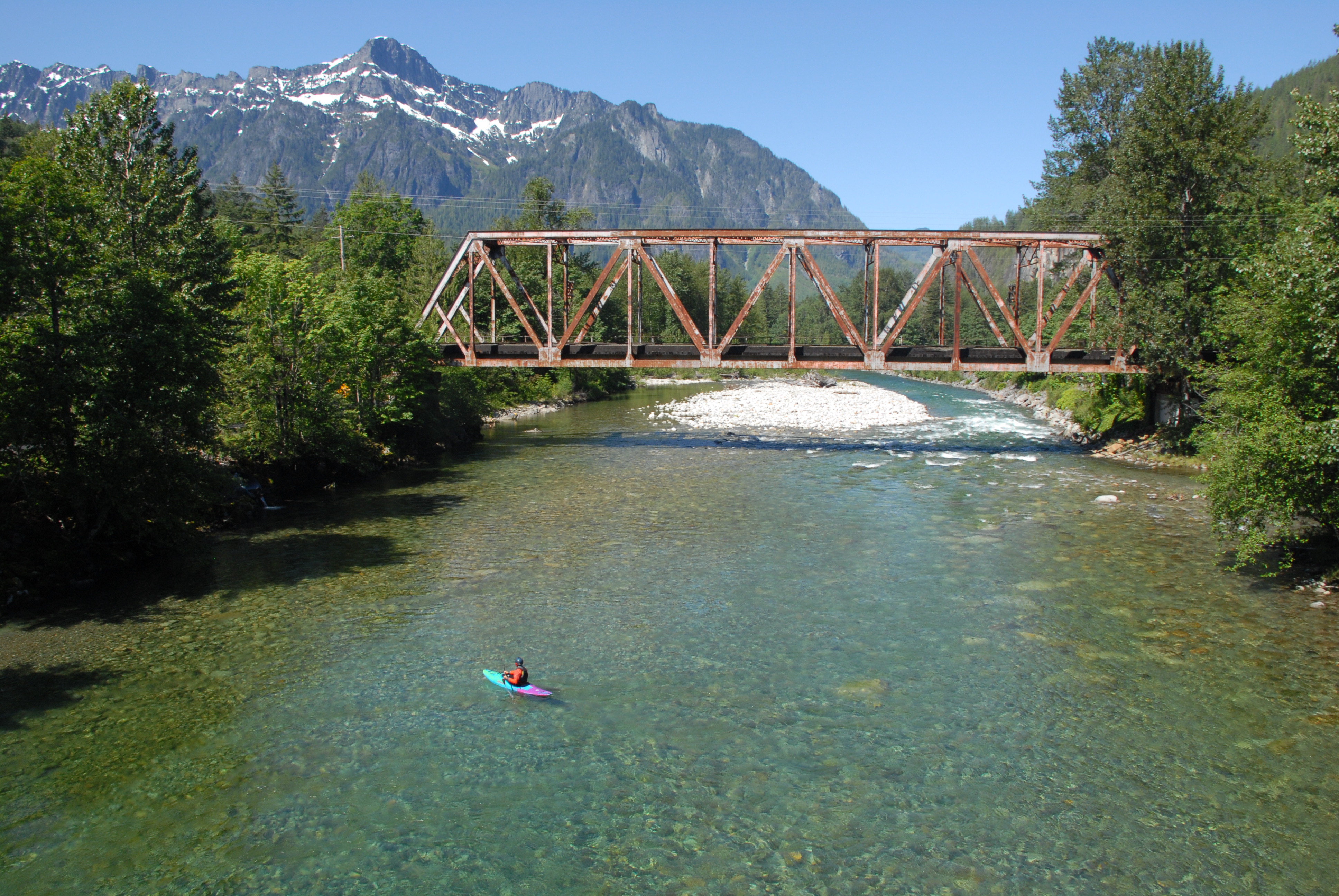 Kayaker launching into the clear North Fork Skykomish River in Index, Washington, with the BNSF Railroad Bridge overhead and forested mountains in the background.